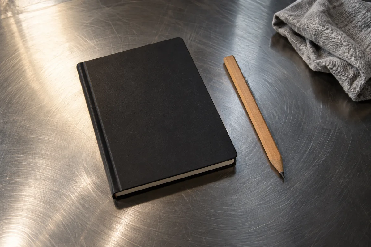 Carnet à couverture noire fermé et crayon de charpentier posés sur une plaque d’acier brut