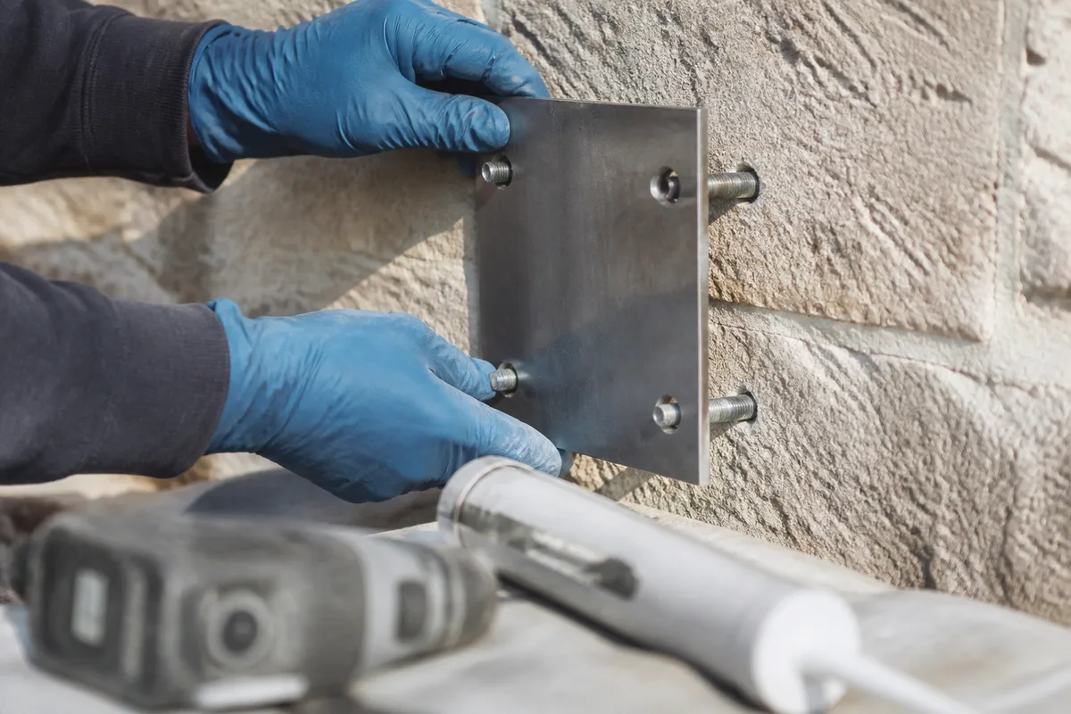 Mains gantées ajustant une platine de fixation de marquise sur un mur en pierre